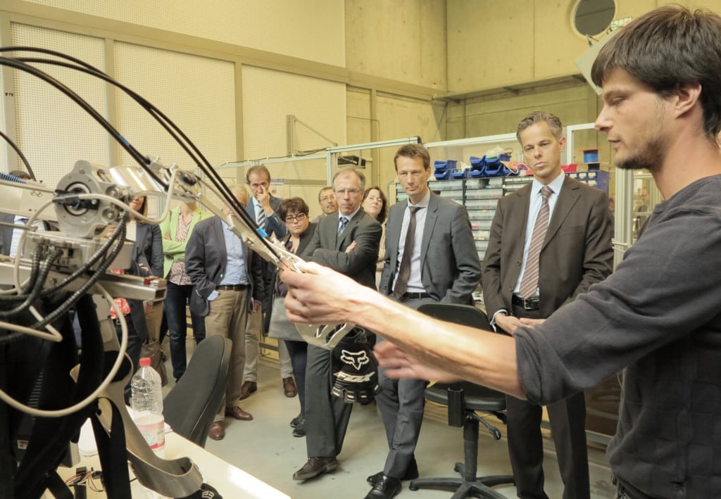 Im Robotiklabor der OTH Regensburg erläuterte der wissenschaftliche Mitarbeiter Benjamin Großmann (rechts) der Delegation der Barmherzigen Brüder die Funktionsweise des Exoskeletts, das an der japanischen Partnerhochschule Tokyo University of Science entwickelt wurde und nun im mit einem an der OTH Regensburg entwickelten Robotersystem gekoppelt wird, um es weiterzuentwickeln. Foto: OTH Regensburg.