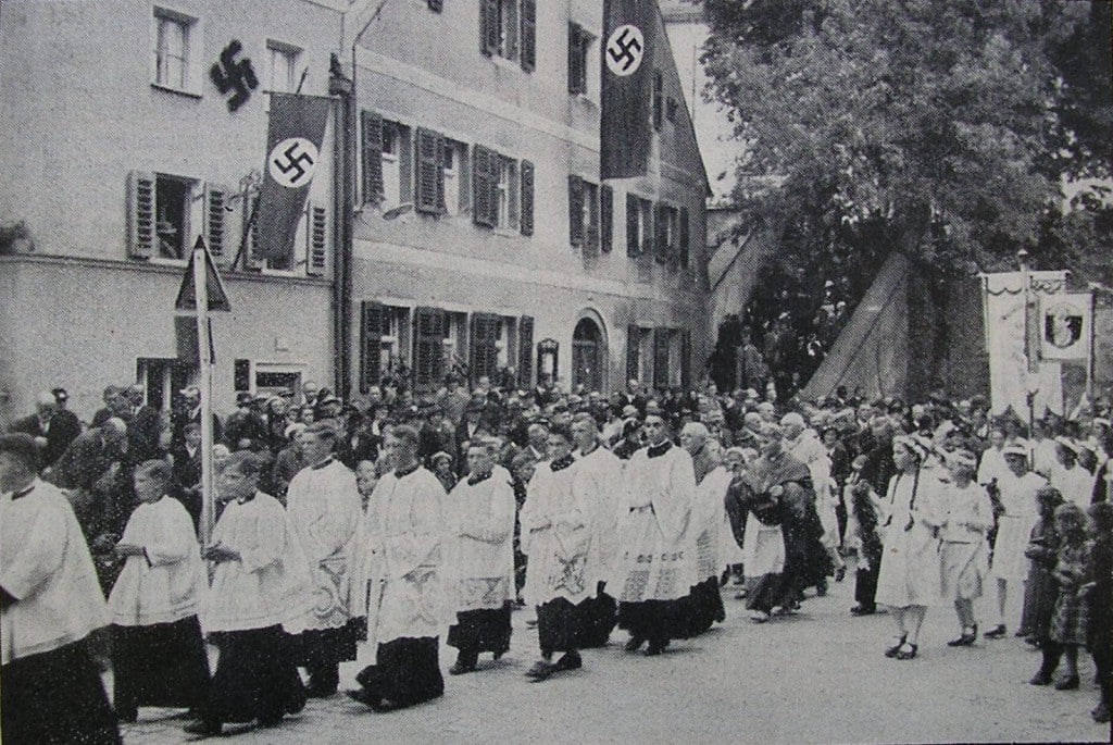 1937: Ein Jahr später feierte man in Deggendorf „600 Jahre Gnadenzeit“ und Bischof Buchberger schritt mit einer feierlichen Prozession durch ein Meer von Hakenkreuzfahnen.